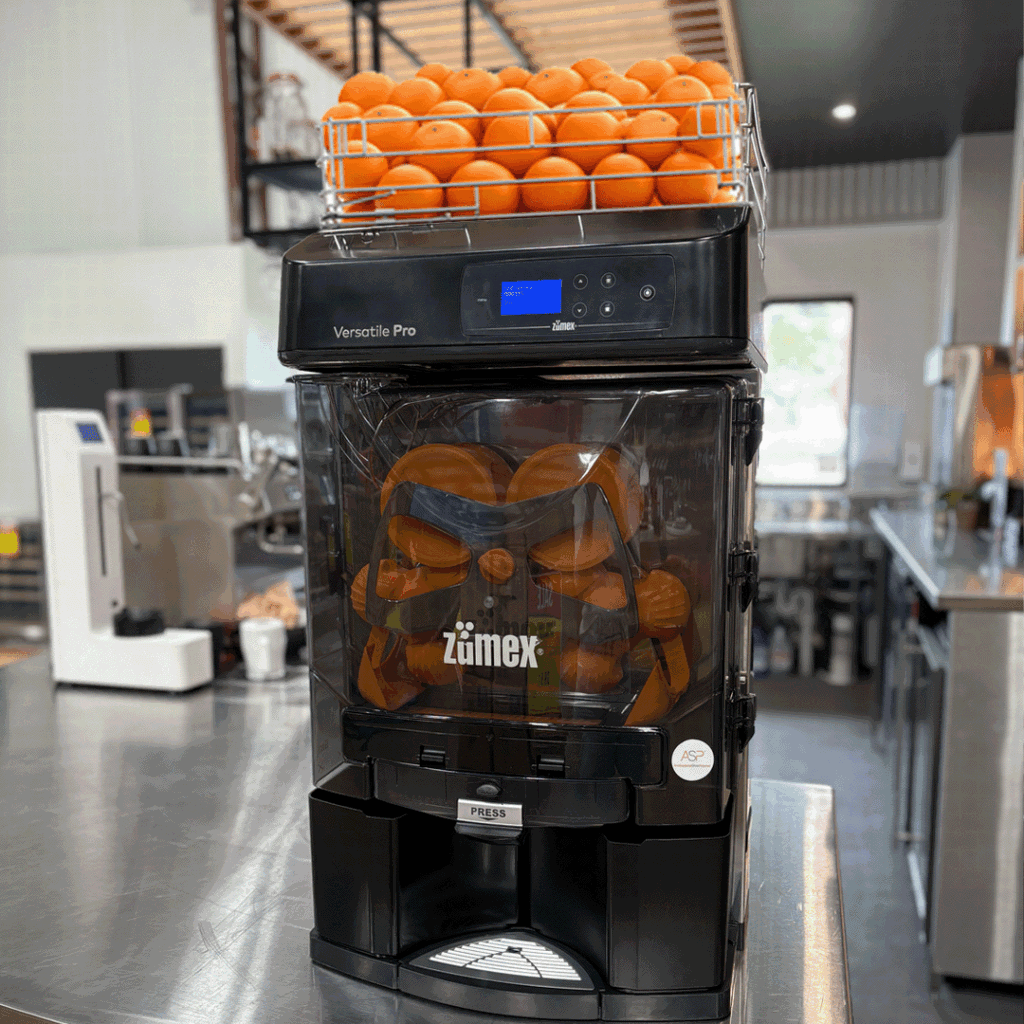 Commercial juicer with orange halves inside the press and a basket of oranges on top in a cafe kitchen setting.