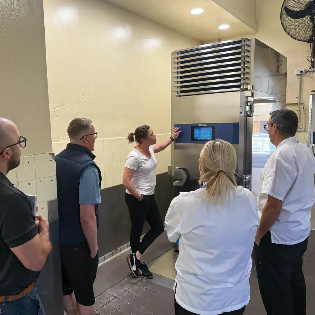 A woman in a white shirt explains an industrial stainless steel unit to four colleagues during a training session in a kitchen area.
