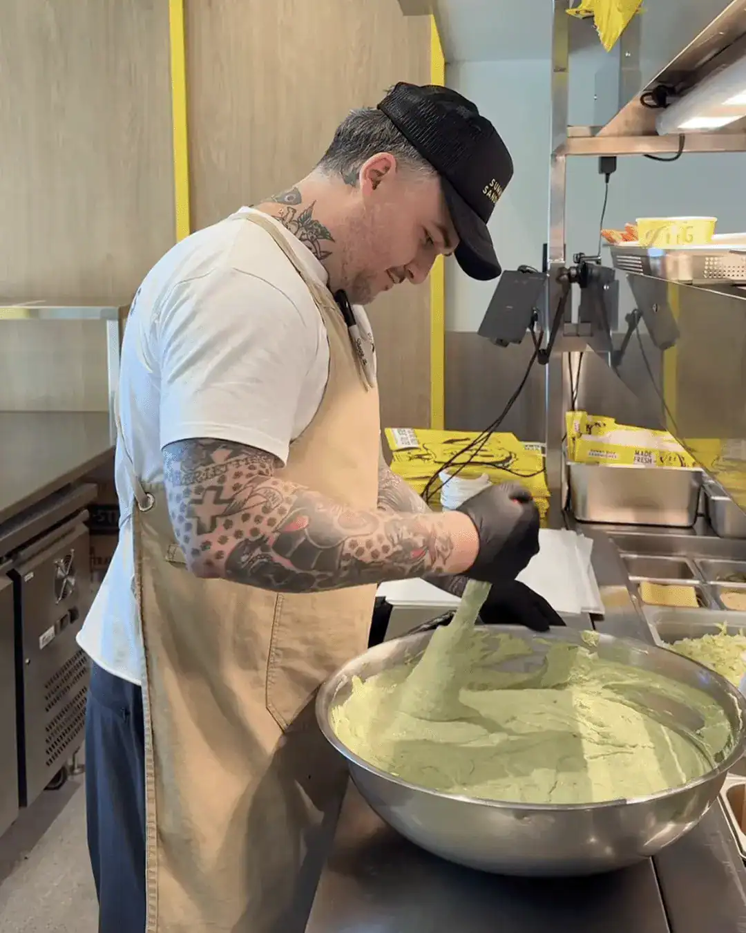 Tattooed chef in a beige apron stirs green pasta dough in a large stainless steel bowl.