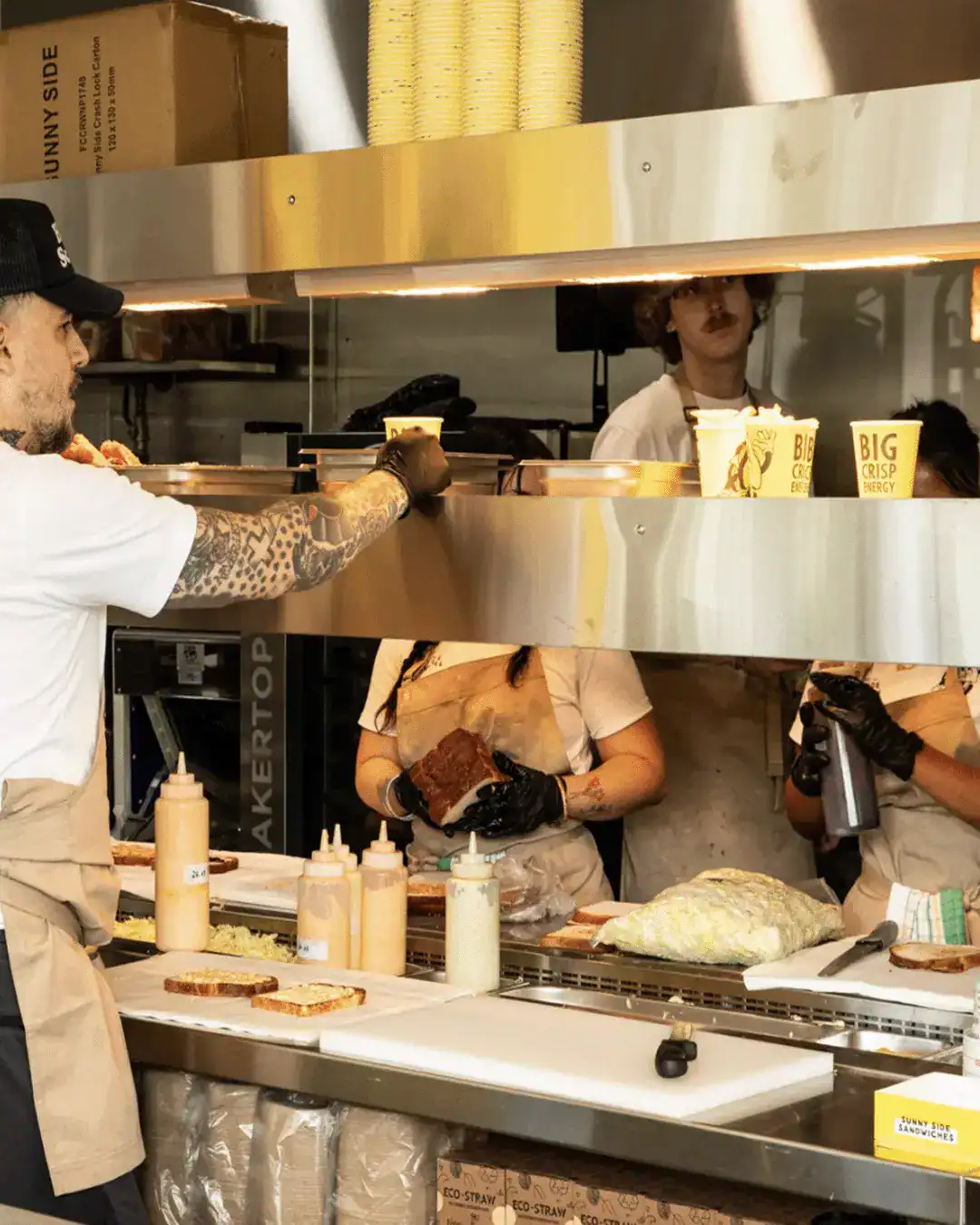 Chef with tattoos handing a sandwich to a customer at a stainless steel counter in a busy kitchen environment.