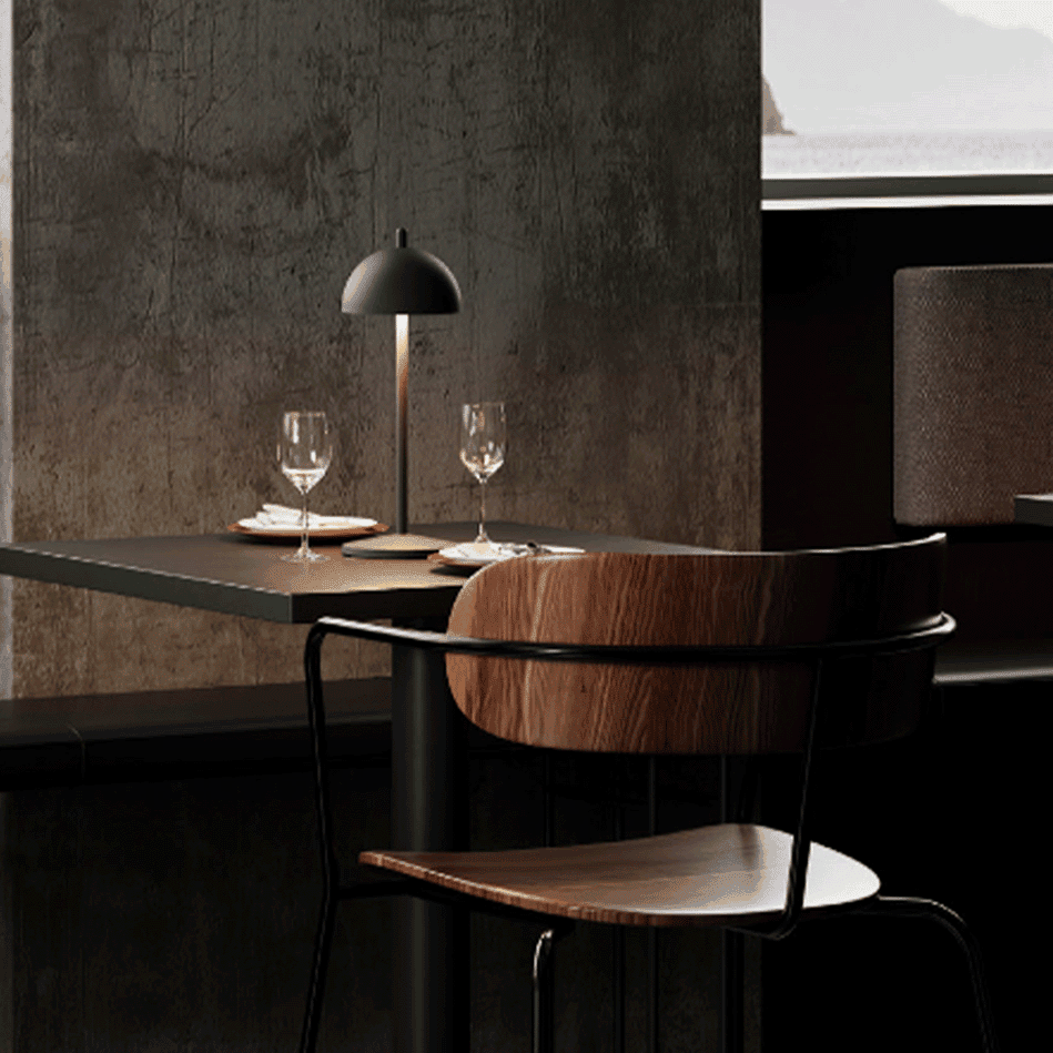 Modern dining table in a dim, stylish restaurant with a black lamp, two wine glasses, and plates set for two against a dark textured wall.