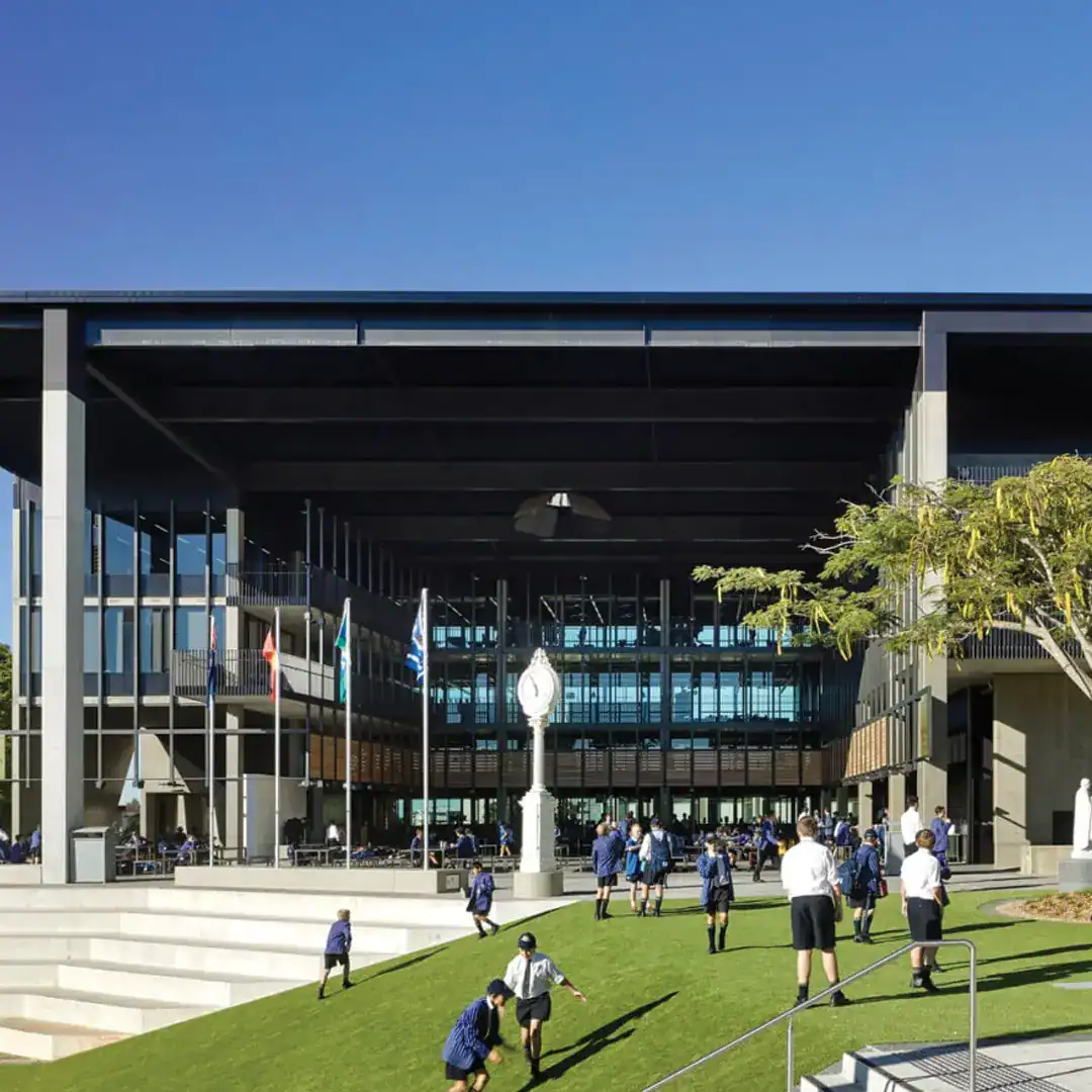 Students in uniforms enjoy the sunny courtyard of a modern campus building with flagpoles and a central clock sculpture on the lawn.