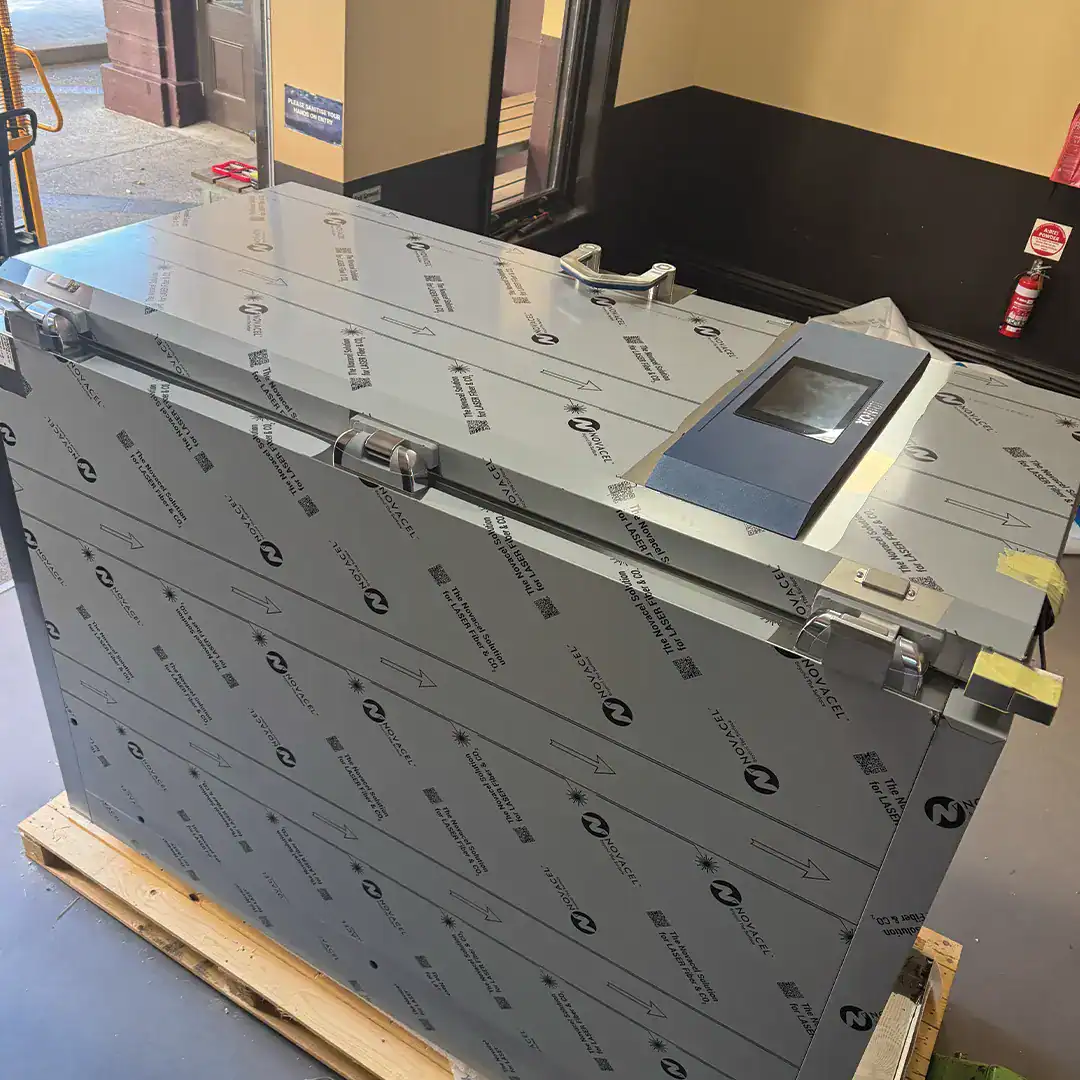 Industrial stainless steel commercial refrigeration unit on a wooden pallet, wrapped in protective film, with a touchscreen control panel nearby a doorway.