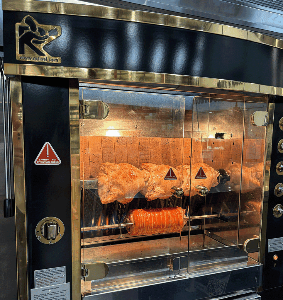 Front view of a black-and-gold Rotisol rotisserie oven with chickens roasting on spits behind glass doors behind metal frame.