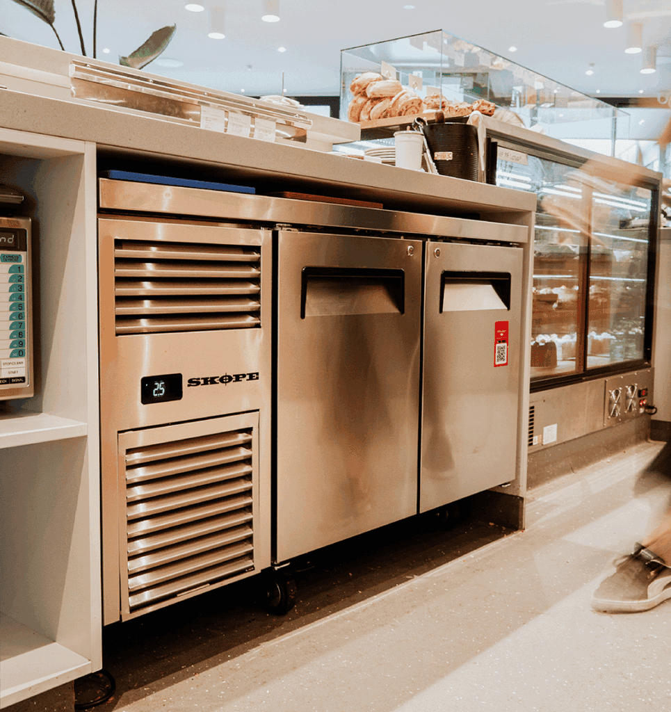 Stainless steel commercial bakery counter with refrigerated drawers and a glass pastry display case on top, bread buns visible behind the glass.