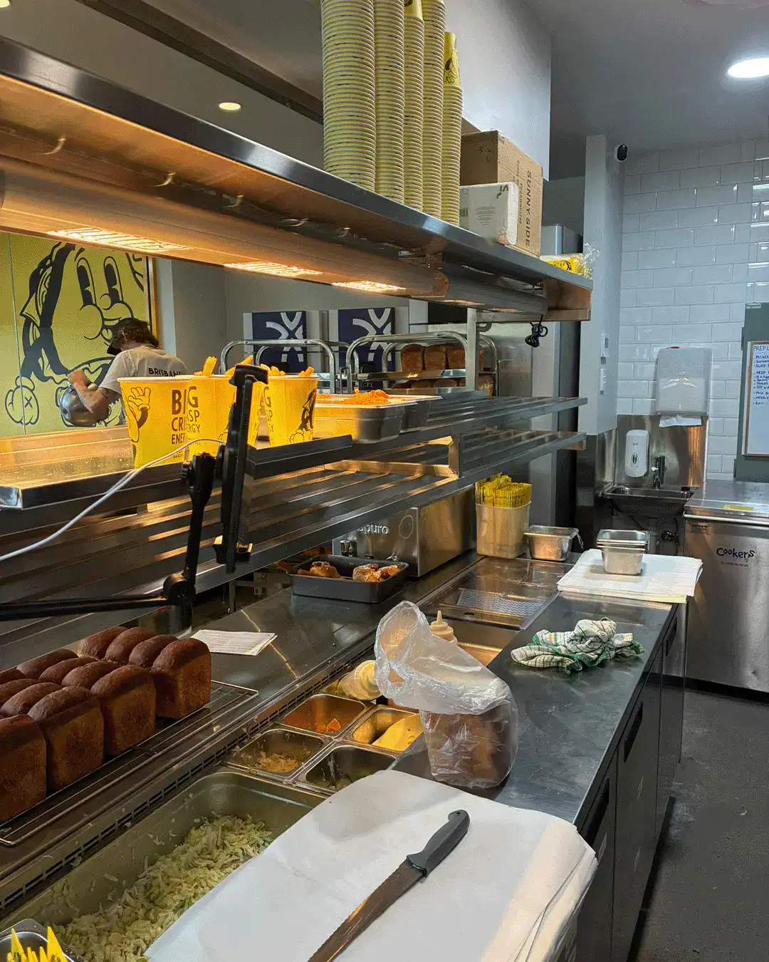 Commercial kitchen prep line with buns, toppings in metal pans, and yellow cups on a stainless counter; a staff member is working in the background under warm lighting.