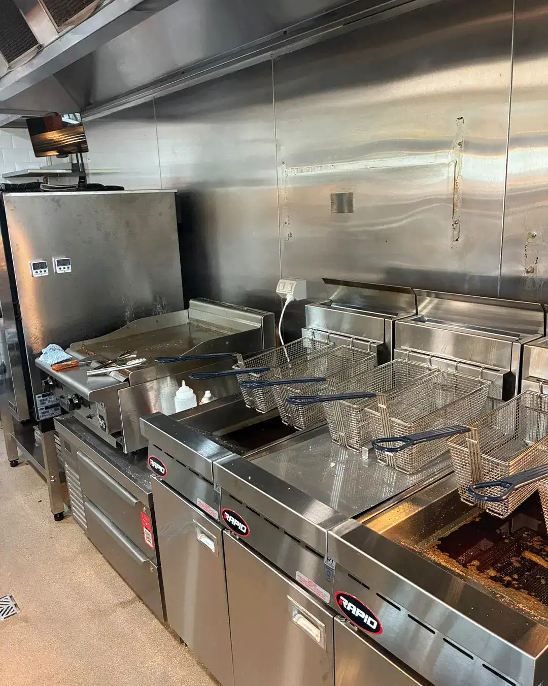 Commercial deep fryer station with multiple wire baskets above hot oil in a stainless steel kitchen setup, ready for frying. Brief cleanup tools nearby.